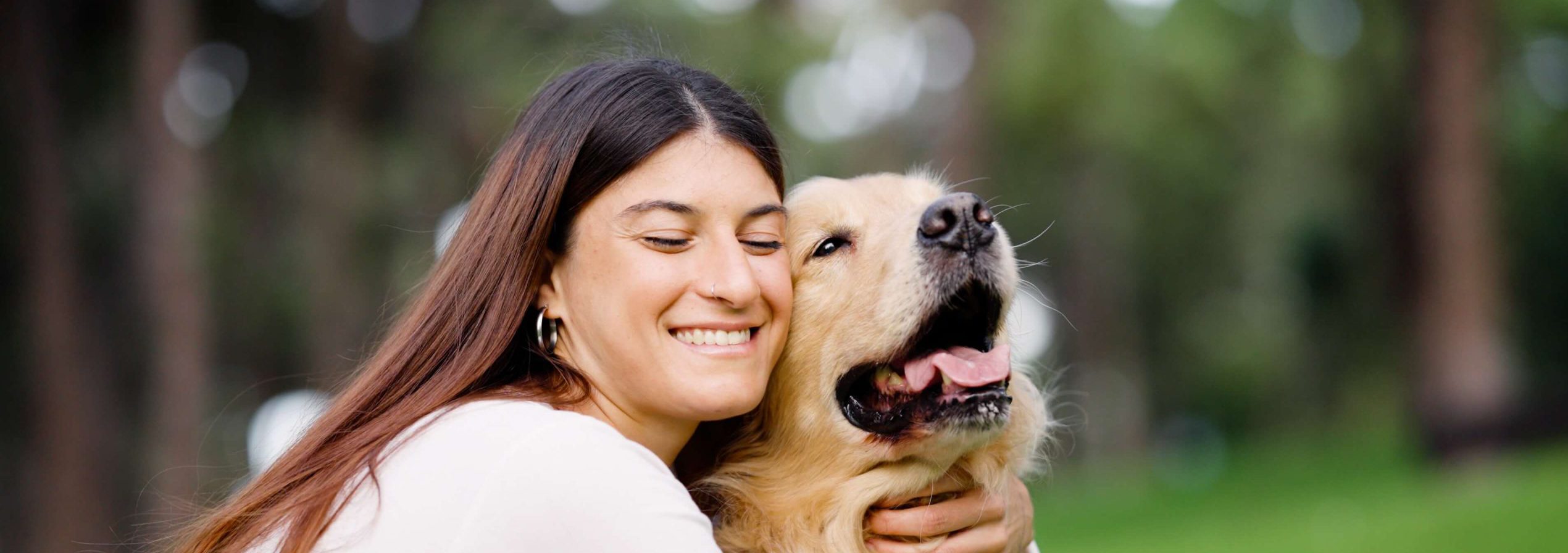 A smiling woman with long hair hugs a happy golden retriever in an orange harness outside, with green grass and trees in the background.